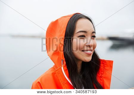 Smiling attractive young asian woman wearing raincoat spending time outdoors walking at the coastland