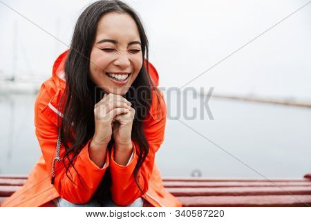 Smiling attractive young asian woman wearing raincoat spending time outdoors walking at the coastland