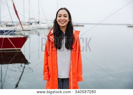Smiling attractive young asian woman wearing raincoat spending time outdoors walking at the coastland