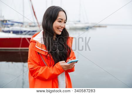 Smiling attractive young asian woman wearing raincoat spending time outdoors walking at the coastland, using mobile phone
