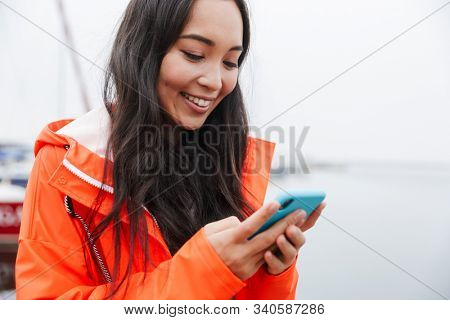 Smiling attractive young asian woman wearing raincoat spending time outdoors walking at the coastland, using mobile phone