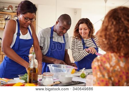 Male And Female Adult Students With Teacher Preparing Ingredients For Dish In Kitchen Cookery Class