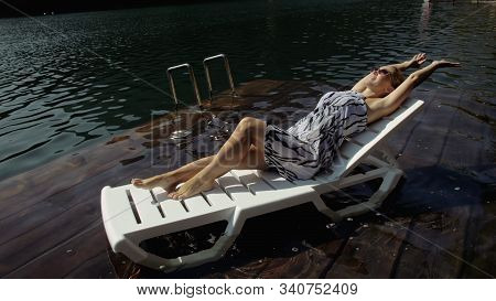 Woman Lie On A Sunbed In Sunglasses And A Boho Silk Shawl. Girl Rest On A Flood Wood Underwater Pier