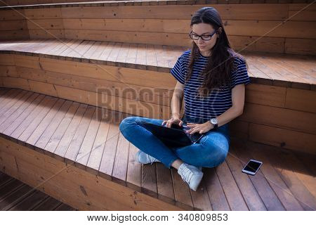A Serious Brunette Student Girl In Glasses, Sits Crosslegged, On Wooden Steps, On A Bench, With An O
