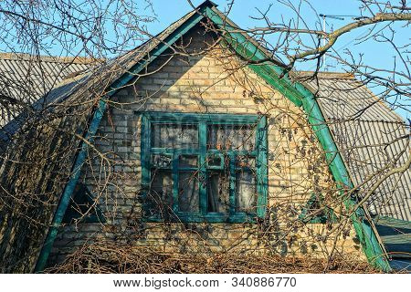 Brown Brick Loft Of A Rural House With A Green Window On A Background Of Gray Branches And A Blue Sk