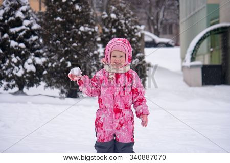 A Happy Smiling Girl In Winter Overalls Is About To Throw A Snowball. Little Girl Joyfully Plays Sno