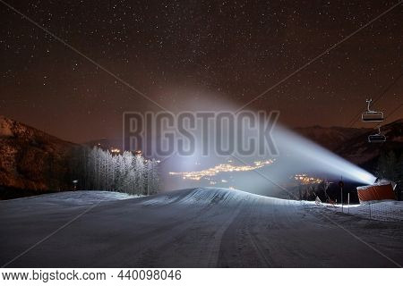 Ski lift in the Alps at night with starry sky above, snow cannon at work, Les Orres 1800 ski resort buildings