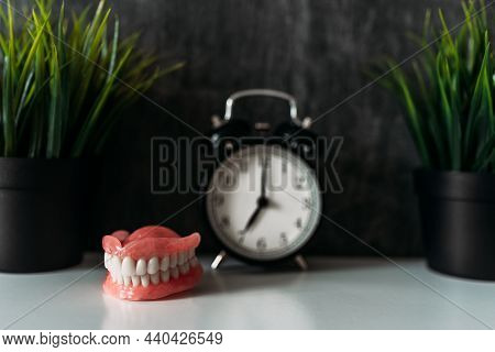 Full Removable Plastic Prosthetic Jaw And Alarm Clock. A Denture Next To The Clock On The Bedside Ta