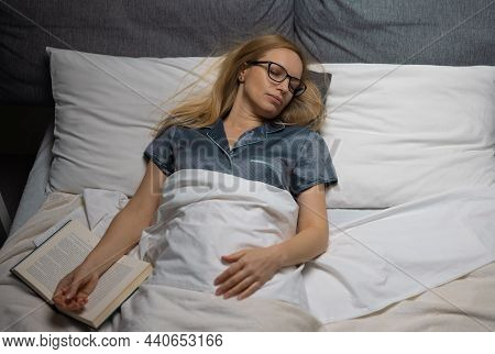 Tired Mature Woman In Eyeglasses And Pajams Falling Asleep While Reading Book In Bed. Female Person 