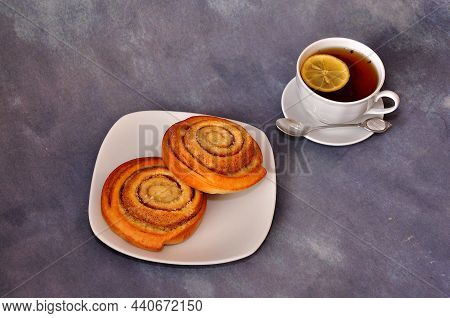 Two Rolled Cinnamon Rolls On A Plate And A Cup Of Black Tea With A Slice Of Lemon On A Gray Abstract