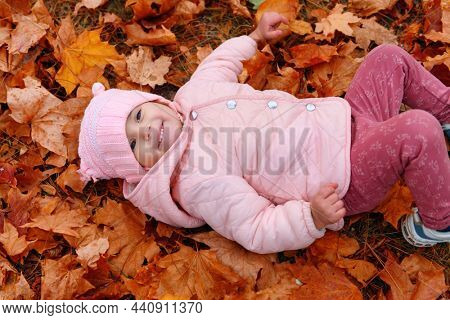 Child girl lying down on fallen leaves in autumn city park. Beautiful nature, trees with yellow leaves.