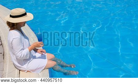 Relaxing Senior Woman Dangling Her Legs In An Outdoor Swimming Pool Holding A Blue Refreshing Cockta
