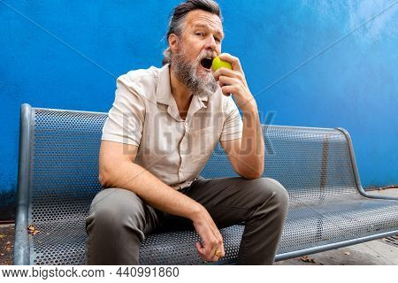 Mature Caucasian Man With Beard Eating An Apple Outdoors Looking At Camera. Blue Background.
