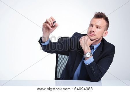 young business man at the desk writing something with his marker on an imaginary screen while holding a hand at his chin in a pensive manner. on a light gray studio background
