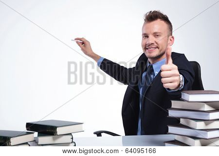 young business man sitting at his desk and showing the thumb up gesture while writing something with chalk on an imaginary screen behind him. on a light studio background
