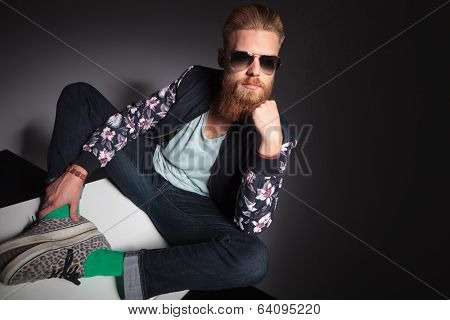 young bearded man looking away while holding his hand at his chin and sitting with his soles together. on a black studio background