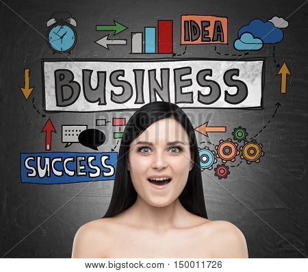Close up of amazed girl with black hair looking at the viewer while standing near blackboard with business sketch on it.