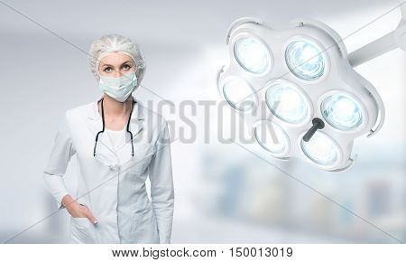 Woman doctor in surgical mask standing near operation room lamp looking down against blurred background. Concept of operation