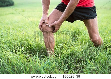 Unrecognizable active senior runner outside in field. Man with injured knee. Close up. Green sunny summer nature.