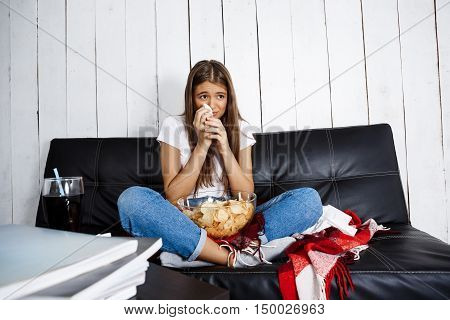 Sad young beautiful girl watching melodrama, crying, sitting on sofa at home. Copy space.