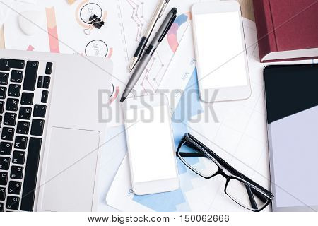 Top view of office desktop with blank white cellphones tablet financial report laptop keyboars supplies and other items. Mock up