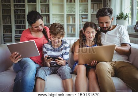Happy family sitting on sofa and using laptop, mobile phone and digital tablet at home