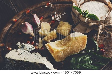 Cheese delikatessen closeup on rustic wood. Wooden round desk with blue cheese roquefort, parmesan, camembert and brie cuts decorated with garlic, pomegranate and basil, french cuisine ingredients