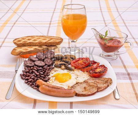 Full English fried breakfast with bacon, egg, sausages, black pudding, mushrooms. Grilled tomatoes and baked beans. Closeup with toasts, coffee, orange juice and home made  red sauce.