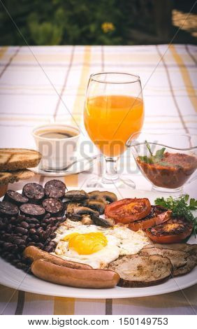 Full English fried breakfast with bacon, egg, sausages, black pudding, mushrooms, grilled tomatoes and baked beans. Closeup with toasts, coffee, orange juice and home made  red sauce.