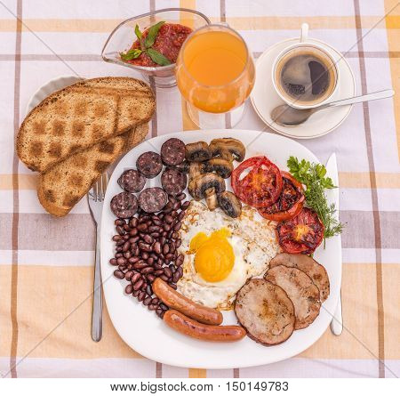Full English fried breakfast with bacon, egg, sausages, black pudding, mushrooms, grilled tomatoes and baked beans. Closeup with toasts, coffee, orange juice and home made  red sauce.