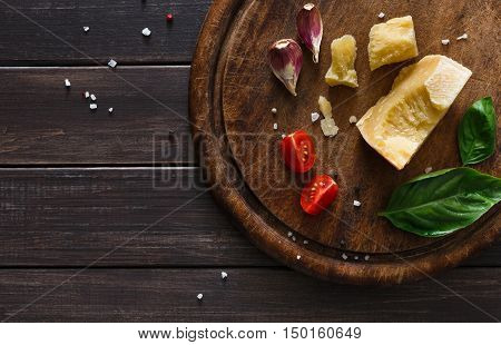 Cheese delikatessen background, closeup on rustic wood. Wooden desk with parmesan, camembert cuts decorated with garlic, tomato and basil, top view with copy space
