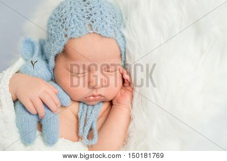 sleepy newborn boy in blue knitted hat with toy on white fluffy blanket