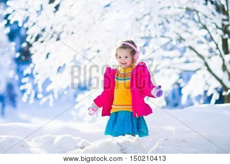 Funny little toddler girl in a colorful knitted hat and warm coat playing with snow. Kids play outdoors in winter. Children having fun at Christmas time. Child enjoying sunny day during Xmas vacation.