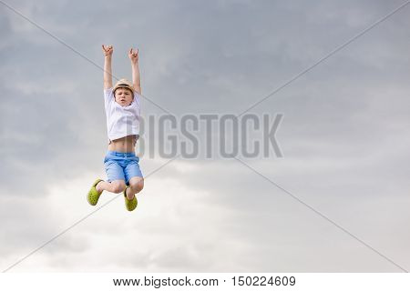 Cute kid boy jumping into cloudy sky - happiness childhood freedom lifestyle concept - smiling boy in straw hat jumping in air over grey rainy sky.