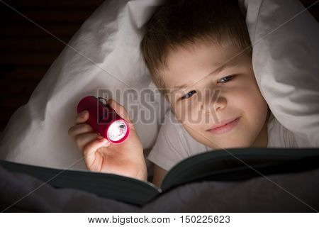 Small kid boy with a smile hiding under a blanket and reading a book with his flashlight in his bedroom at night. Enjoying learning and books.