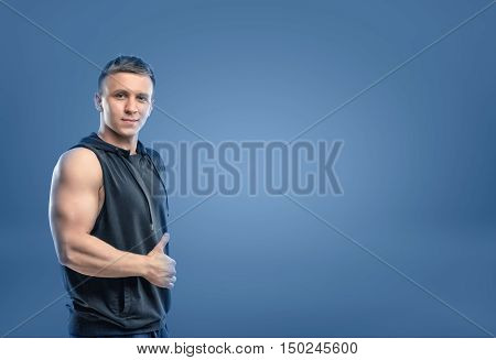 Croppeed portrait of muscled young man showing his bicep and thumbs up on blue background. Self-improvement. Athlete and bodybuilder. Workout and fitness.