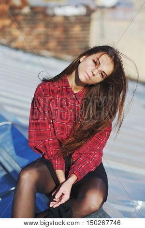 the pretty Young girl in a red plaid blouse and black short skirt sitting on the roof of the house. The wind ruffled her hair.