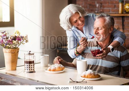 Spend time with pleasure. Smiling delighted elderly woman feeding her lovely husband with berries while enjoying the celebration and sitting in the kitchen.