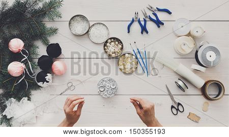 Making handmade christmas balls. Woman's leisure, tools for creating holiday decorations and garland. Top view of white wooden table with female hands holding various trinkets, decor accessories