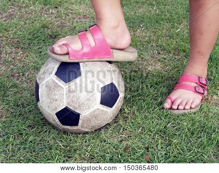 close up leg of Asian little girl wear pink sandal stand on soccer ball on green grass field, outdoor sport fun for children