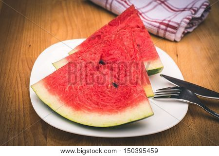 Watermelon slices on the white porcelain plate served with cutlery and napkin straw-coloured wooden background