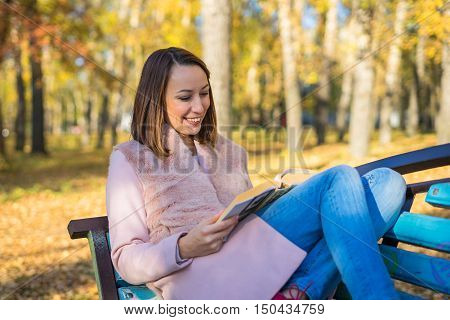 Girl sits on a park bench and reading a book sunny autumn day in October. Yellow leaves underfoot and the trees.