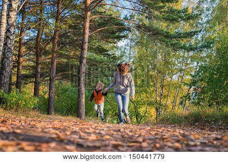Mother and son running on path in autumn forest. Family at nature autumn bright sunny day fallen yellow leaves underfoot.