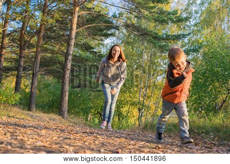 Mother and son running on path in autumn forest. Family at nature autumn bright sunny day fallen yellow leaves underfoot.