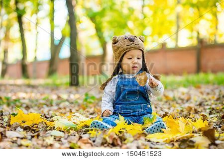 Happy happy baby sitting in the park with yellow leaves in autumn in bear cap. In autumn park kid sitting in yellow leaves. Cheerful baby sitting in the park in bear cap. Warm autumn day in the park.