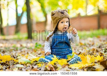 Happy happy baby sitting in the park with yellow leaves in autumn in bear cap. In autumn park kid sitting in yellow leaves. Cheerful baby sitting in the park in bear cap. Warm autumn day in the park.