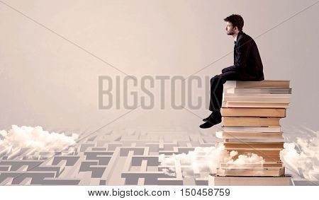 A serious businessman in suit sitting on a pile of giant books in front of a grey wall with clouds, labirynth