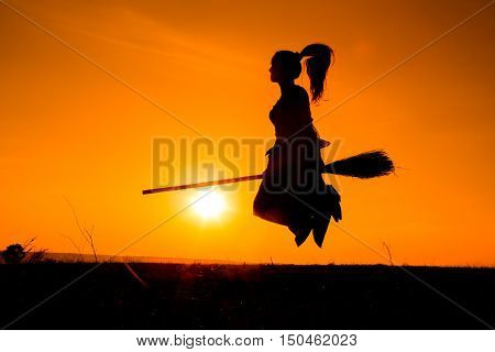Silhouette of young witch flying on the broomstick against sunset