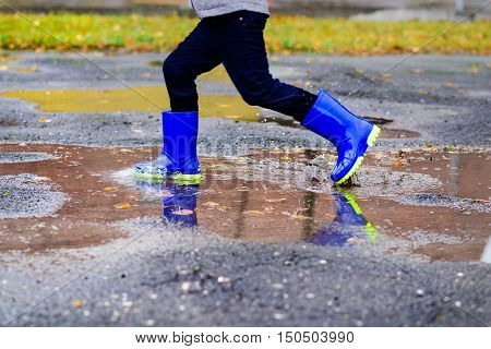 Boy In Rubber Blue Rainboots Jumping To Dirty Puddle
