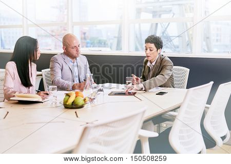 Business meeting between three executive employees, two female and one male, taking place in a birght conference table early in the morning to prepare for the week ahead.